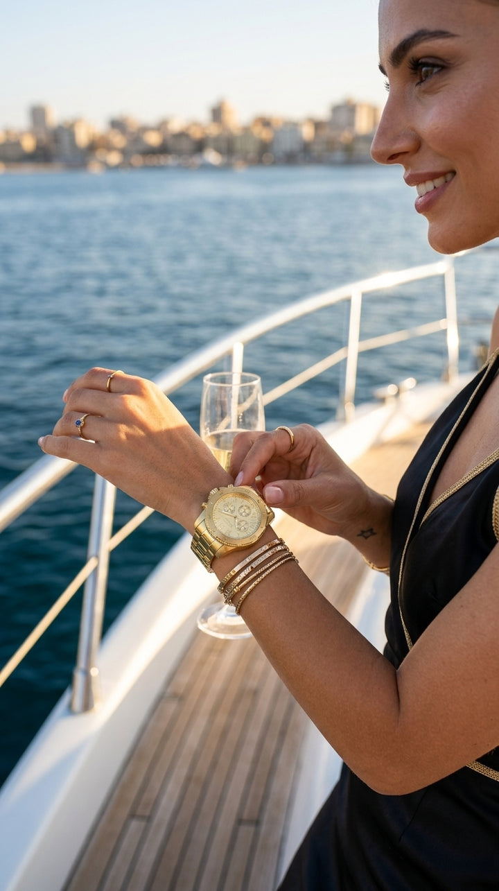 A woman on a yacht wearing a gold-tone watch with a cream dial, adjusting it while holding a glass of champagne. The background shows the blue water and a distant city skyline under warm sunlight.